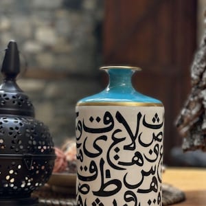 Può includere: Vaso in ceramica con base bianca, decorato con calligrafia nera. La parte superiore del vaso è turchese, con una fascia dorata che la separa dal corpo bianco. A sinistra c'è una lanterna nera decorativa.
