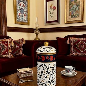 May include: A cylindrical ceramic jar with a lid, featuring black Arabic calligraphy on a white background, a dark blue band with red floral motifs, and gold trim. The jar is placed on a wooden table alongside a small patterned cup and saucer.