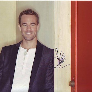 May include: A man wearing a dark blue blazer and a white t-shirt smiles at the camera. He is standing in front of a red door with a silver door handle and the number "23" on it.