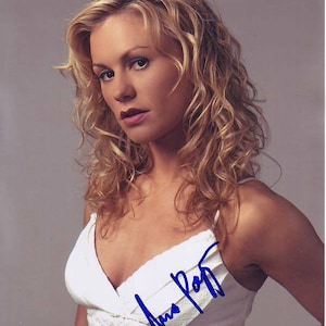 Puede incluir: Una mujer con el pelo largo, rubio y rizado lleva un top blanco sin mangas con un patrón de encaje. El top tiene un escote en V profundo y la firma de la mujer está escrita en tinta azul en la parte delantera del top.