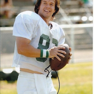 May include: A man wearing a white jersey with green numbers and a white wristband holds a brown football. He is wearing white pants and has long brown hair.