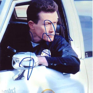 May include: A man in a police uniform sits in a white car with the words "protect and to serve" and the City of Los Angeles seal on the side. He is looking down and has signed his name in blue ink on the side of the car.