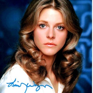 May include: A close-up portrait of a woman with long, flowing brown hair. She is wearing a white shirt and has a serious expression on her face. The image is signed "Jamie Lee Curtis".