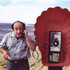 Może przedstawiać: Mężczyzna w jasnoniebieskiej koszuli rozmawia przez czerwony telefon publiczny w kształcie muszli. Telefon publiczny znajduje się na plaży z niebieskim oceanem w tle.