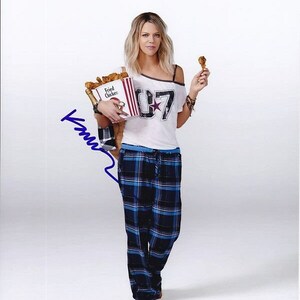 May include: A woman wearing a white t-shirt with the number "07" and a star on it, blue and black plaid pajama pants, and holding a bucket of fried chicken and a chicken leg. The woman is smiling and looking at the camera.