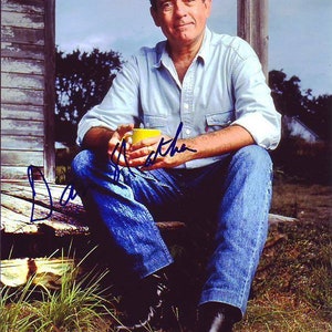Pode incluir: Um homem vestindo uma camisa azul claro abotoada e jeans azul senta-se em uma varanda de madeira com uma caneca amarela na mão. Ele está olhando para o lado e sorrindo. O fundo é uma imagem desfocada de uma casa e árvores.