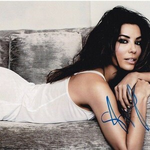 Puede incluir: Una mujer con el pelo largo y oscuro está tumbada en un sofá gris. Lleva un top blanco sin mangas y tiene una firma azul en el brazo.