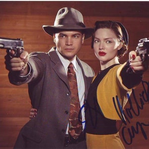 Puede incluir: Un hombre y una mujer con ropa vintage, ambos sosteniendo armas de fuego, con un fondo de madera. La mujer lleva una chaqueta amarilla y un vestido negro. El hombre lleva un traje gris y un sombrero fedora. La firma de la mujer es visible en la esquina inferior derecha.