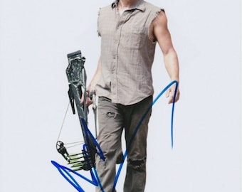 May include: A man with brown hair and a beard is wearing a gray sleeveless shirt and blue jeans. He is holding a black crossbow with green arrows. The man is looking at the camera and has a serious expression on his face. He has signed his name in blue ink.