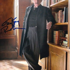 May include: A man in a black coat and striped pants stands next to a wooden bookshelf filled with books. He is looking at the camera and has a mustache. The man's signature is visible in the lower left corner of the image.