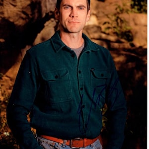 May include: A man wearing a dark green button-down shirt with a brown belt and blue jeans. He is standing in front of a rock wall.