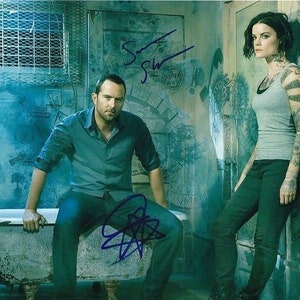 Puede incluir: Un hombre y una mujer están de pie en una habitación con una pared de azulejos azules y blancos. El hombre lleva una camisa azul y pantalones vaqueros. La mujer lleva un top gris sin mangas y pantalones negros. Hay una bañera en la habitación.