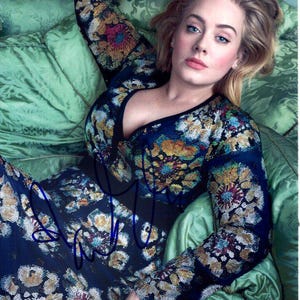 Puede incluir: Una mujer con un vestido azul oscuro de manga larga con un estampado floral en tonos amarillos, naranjas y blancos. El vestido tiene un escote pronunciado y está firmado. Está tumbada sobre una superficie verde estampada.