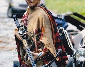 May include: A man with short brown hair is holding a crossbow. He is wearing a brown and red patterned poncho. The crossbow is black and has a blue string. The man is standing in front of a motorcycle.