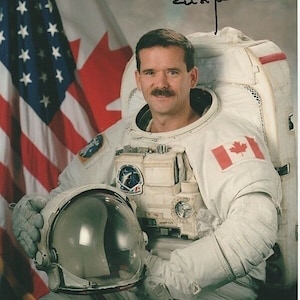 Puede incluir: Un hombre con un traje espacial blanco con un parche de la bandera canadiense en el hombro izquierdo. Está sosteniendo un casco blanco en su mano izquierda. Está de pie frente a una bandera estadounidense y una bandera canadiense.