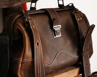 May include: A brown leather satchel with white stitching and a metal clasp. The bag has a top handle and a detachable shoulder strap. The leather has a distressed look, and the bag appears to be sitting on a wooden shelf.