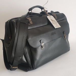 May include: A black leather briefcase with a top handle and a detachable shoulder strap. The bag features metal clasps, a flap closure, and the initials "JSG" embossed on the front. A tag hangs from the handle.