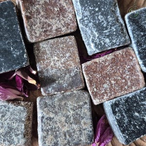 May include: A close-up of several square, textured soap bars in shades of brown, grey, and black. The soaps are arranged closely together, with a few dried purple flower petals scattered around them.