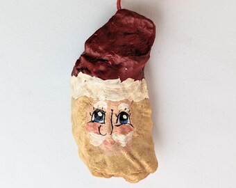 May include: A hand-painted oyster shell ornament depicting Santa Claus with a red hat, white beard, and blue eyes. The ornament has a red string for hanging.