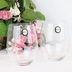 May include: Two clear, stemless wine glasses with the "RIEDEL" logo on the front. The glasses are oval-shaped and sit on a white surface. Pink flowers and greenery are visible behind the glasses, adding a touch of color and elegance.