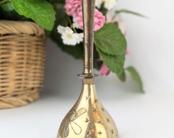 May include: A small, brass bell with a star-shaped design and a dragonfly etched on the side. The bell has a dark brown handle.