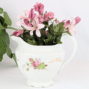 May include: A white porcelain pitcher with a floral design and a pink flower arrangement inside. The pitcher has a scalloped edge and a handle.
