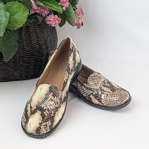 May include: A pair of slip-on shoes with a snakeskin pattern in shades of tan, brown, and black. The shoes have a rounded toe and a black sole. The shoes are displayed on a white surface.