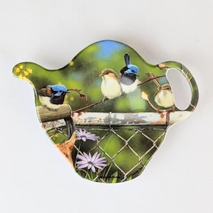 May include: A ceramic teapot-shaped tea bag holder with a green, blue, and yellow design featuring four birds perched on a fence. The holder has a handle and a spout.