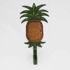 May include: A metal pineapple-shaped hook with a green and brown painted design. The hook is designed to hang items on a wall.