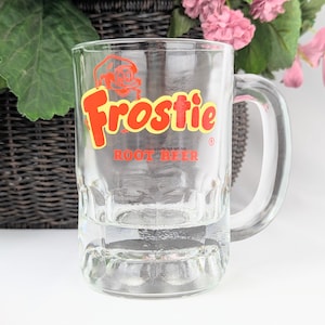 May include: Clear glass Frostie Root Beer mug with a large handle. The mug features the Frostie logo in red and yellow. The mug is rectangular with a thick base and a clear handle.