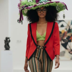 Könnte beinhalten: Ein Model trägt einen roten Blazer mit schwarzem Besatz, ein grünes Strickoberteil und eine gestreifte Hose. Das Model trägt außerdem einen großen, dekorativen Hut mit grünen und lila Akzenten und hängenden Verzierungen. Die Brille hat einen dicken, dunklen Rahmen.