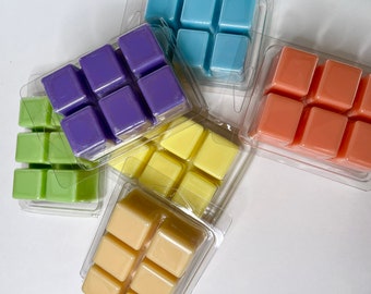 May include: An overhead shot of several clear plastic clamshells, each containing six colorful wax melt cubes. The colors include purple, light blue, orange, yellow, green, and cream. The wax melts are arranged on a white surface.