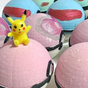 May include: A collection of colorful bath bombs, some with a design resembling a Pokeball. A yellow Pikachu figurine sits atop one of the pink bath bombs. The bath bombs are various shades of blue, pink, and green.