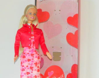 May include: A Barbie doll wearing a red jacket over a pink dress with heart patterns. The doll has blonde hair and is standing in front of a box with heart-shaped decorations and the word "Barbie" on top.