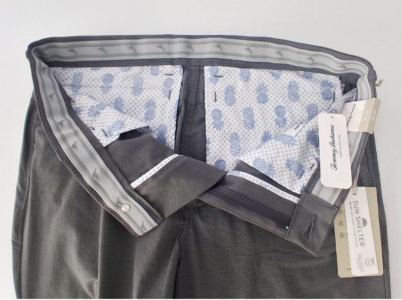 K&ouml;nnte beinhalten: Dunkelgraue Hose mit einem Bund, der ein wiederkehrendes Marlin-Logo-Muster aufweist. Das Innenfutter zeigt einen hellblauen Ananasdruck auf wei&szlig;em Grund. Die angebrachten Etiketten enthalten den Markennamen Tommy Bahama und den Text Sun Shelter.