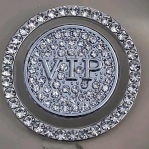 May include: A silver-coloured, circular accessory with a central disc bearing the letters "VIP" in raised lettering. The disc is encrusted with numerous small, clear crystals, surrounded by a ring of larger, similarly sparkling crystals.
