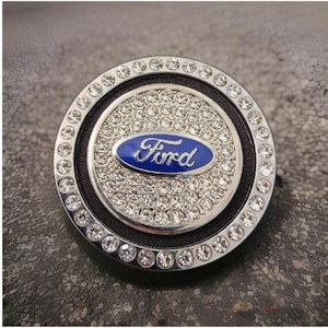 May include: A circular emblem with the Ford logo in blue, set against a silver backdrop, and encircled by sparkling rhinestones. The emblem is mounted on a black base with a silver outer ring, a glamorous car accessory.