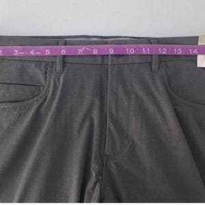 K&ouml;nnte beinhalten: Dunkelgraue Hose aus strukturiertem Stoff mit Knopfverschluss und G&uuml;rtelschlaufen. Ein rosa Ma&szlig;band liegt &uuml;ber dem Bund und zeigt ein Ma&szlig; von etwa 43 cm. Ein wei&szlig;es Etikett ist am Hosenbund befestigt.
