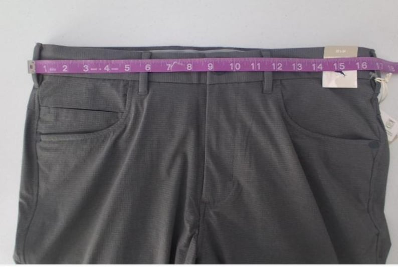 K&ouml;nnte beinhalten: Dunkelgraue Hose aus strukturiertem Stoff mit Knopfverschluss und G&uuml;rtelschlaufen. Ein rosa Ma&szlig;band liegt &uuml;ber dem Bund und zeigt ein Ma&szlig; von etwa 43 cm. Ein wei&szlig;es Etikett ist am Hosenbund befestigt.