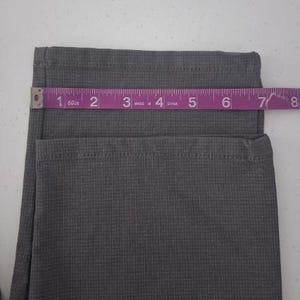 K&ouml;nnte beinhalten: Eine graue Hose mit einem dezenten Karomuster und einem ges&auml;umten Beinabschluss. Ein violettes Ma&szlig;band liegt horizontal &uuml;ber dem Stoff und zeigt eine Breite von etwa 19 cm an der Bein&ouml;ffnung an.