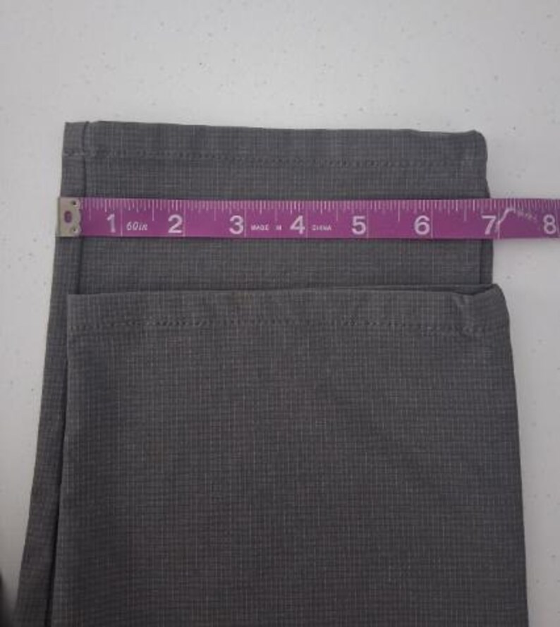 K&ouml;nnte beinhalten: Eine graue Hose mit einem dezenten Karomuster und einem ges&auml;umten Beinabschluss. Ein violettes Ma&szlig;band liegt horizontal &uuml;ber dem Stoff und zeigt eine Breite von etwa 19 cm an der Bein&ouml;ffnung an.
