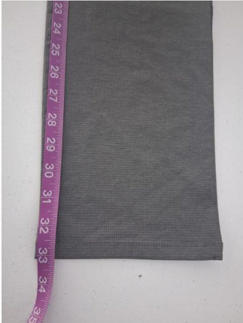 K&ouml;nnte beinhalten: Ein graues Stoffhosenbein mit feiner horizontaler Rippenstruktur und einem gen&auml;hten Saum. Ein violettes Ma&szlig;band ist vertikal an der Seite angebracht und zeigt eine L&auml;ngenmessung, die von 58 cm bis 89 cm reicht.