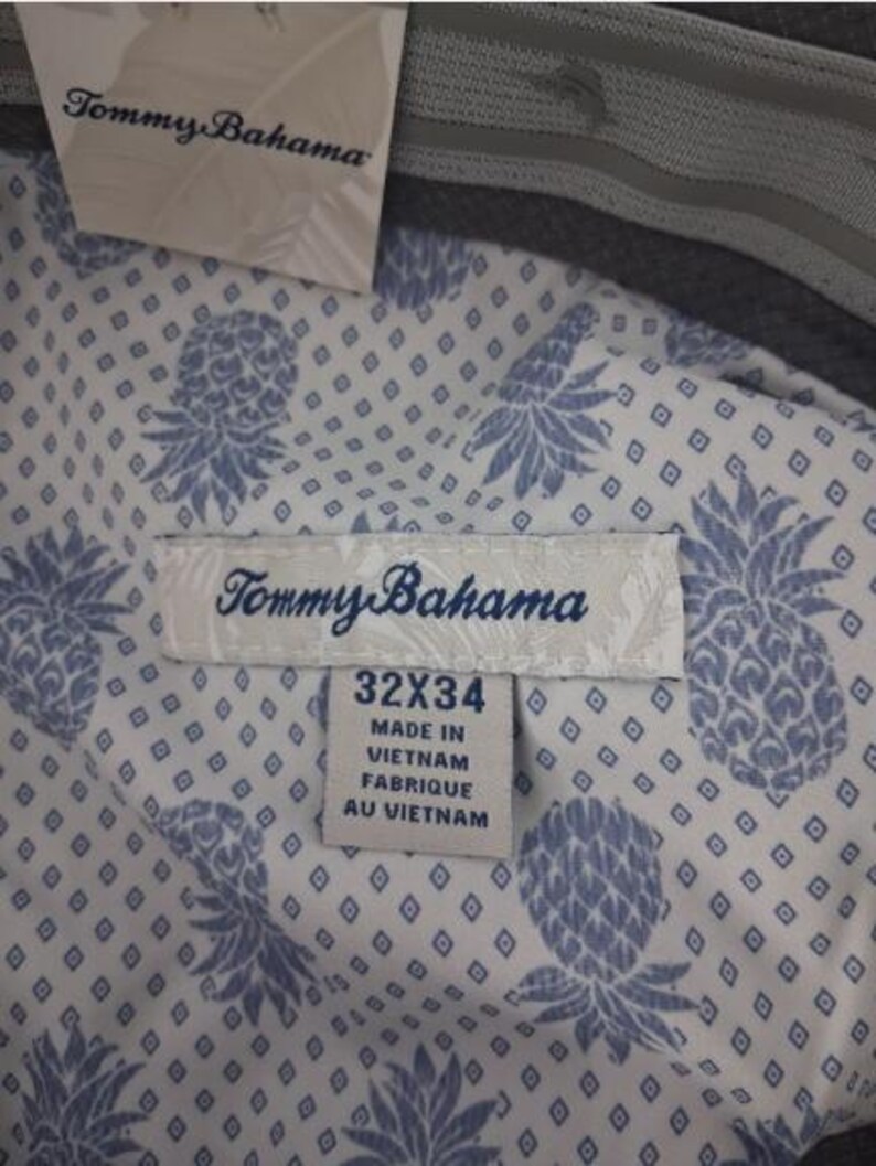 K&ouml;nnte beinhalten: Eine Hose mit wei&szlig;em Innenfutter, das mit einem blauen Ananas- und Rautenmuster bedruckt ist. Ein Tommy Bahama Markenschild ist befestigt. Das Gr&ouml;&szlig;enetikett zeigt 32x34 und gibt an, dass das Kleidungsst&uuml;ck in Vietnam hergestellt wurde. Der Bund ist grau.