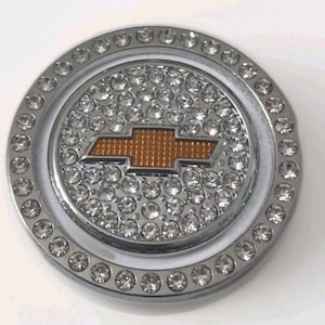 May include: A circular chrome emblem featuring a central gold-toned Chevrolet bowtie logo. The emblem is surrounded by a border of clear, faceted rhinestones, with additional rhinestones covering the entire circular surface around the central logo.