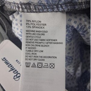 K&ouml;nnte beinhalten: Ein wei&szlig;es Pflegeetikett mit schwarzem Text, der 78% Nylon, 9% Polyester und 13% Elasthan sowie Waschanleitungen und Symbole auflistet. Das Etikett ist an einem grauen Stoff mit blau-wei&szlig;em Blumenmuster und einem silbernen Knopf befestigt.