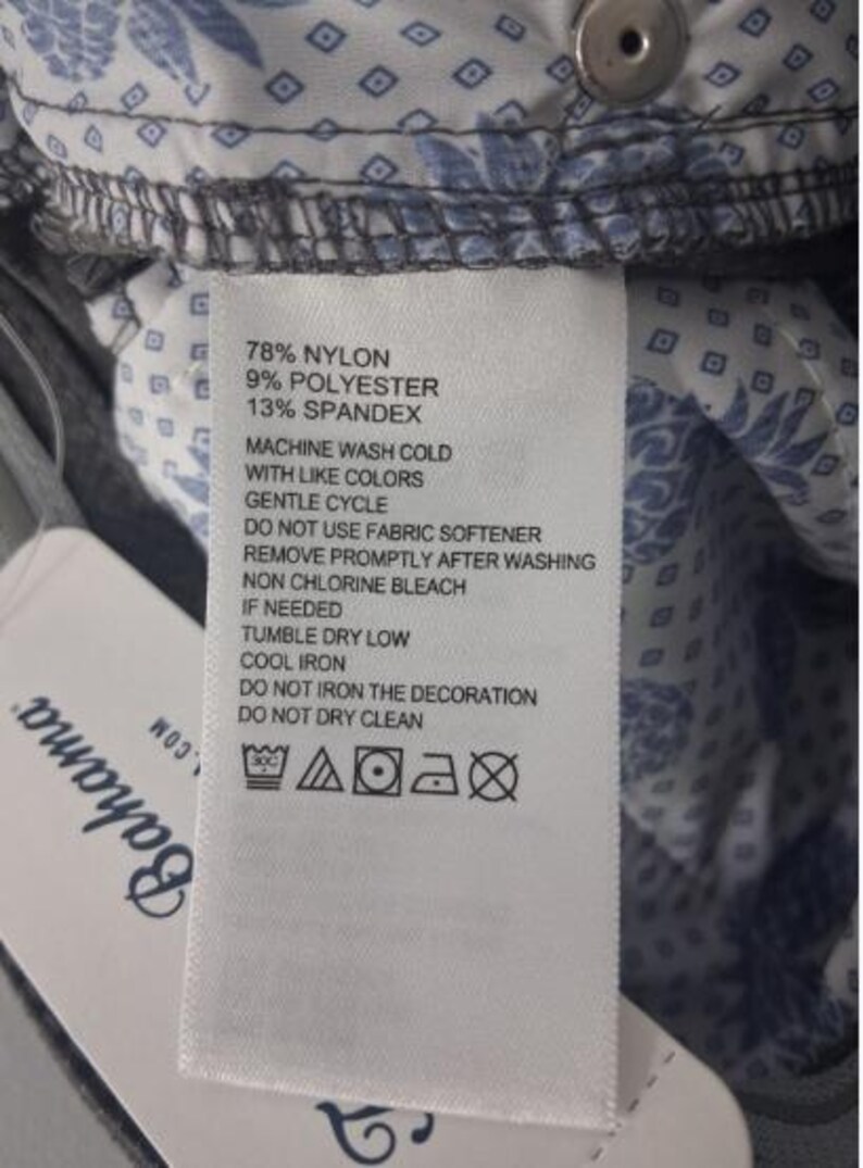 K&ouml;nnte beinhalten: Ein wei&szlig;es Pflegeetikett mit schwarzem Text, der 78% Nylon, 9% Polyester und 13% Elasthan sowie Waschanleitungen und Symbole auflistet. Das Etikett ist an einem grauen Stoff mit blau-wei&szlig;em Blumenmuster und einem silbernen Knopf befestigt.