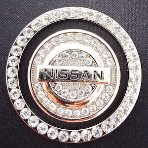 May include: A silver-toned car emblem with the brand name "NISSAN" in a rectangular bar. The emblem is circular, with multiple concentric rings and embedded clear crystals. The background is black.