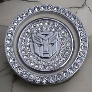 May include: A circular, silver-coloured emblem with the Autobot symbol in the centre, encircled by a ring of clear, sparkling crystals. The emblem has a polished finish, and the crystals add a touch of glamour to the design.