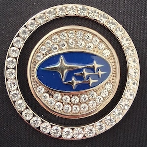 May include: A circular, silver-coloured emblem with a blue oval centre displaying a star cluster design. The emblem is surrounded by two rings of clear, sparkling rhinestones, creating a glamorous and eye-catching accessory.