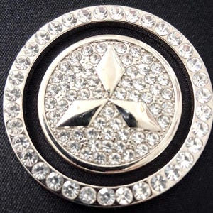 May include: A silver emblem featuring a central logo surrounded by a circle of clear crystals. The logo has a three-diamond design, also encrusted with crystals. The emblem is set against a dark background.